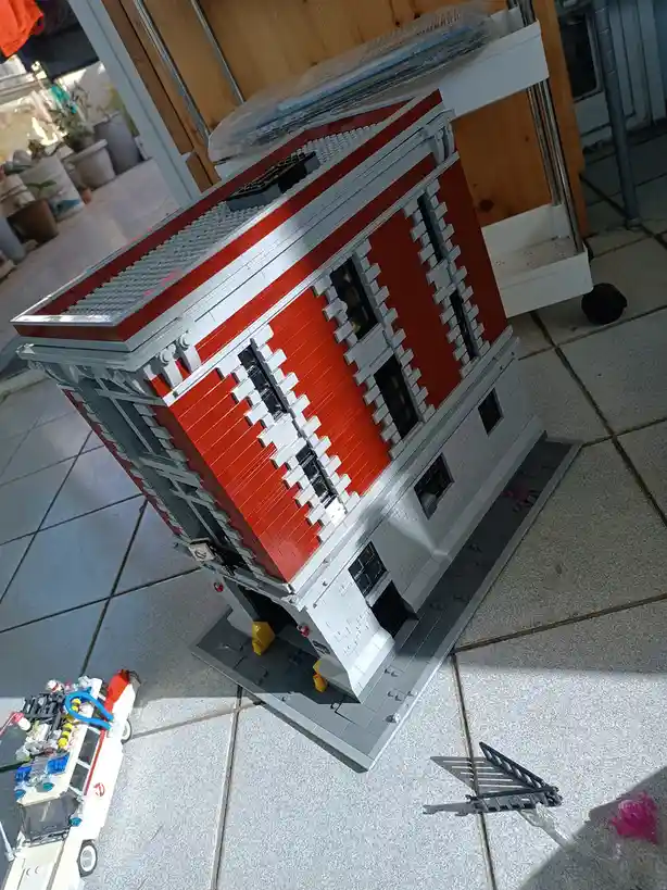 A large, detailed LEGO model of the Ghostbusters firehouse building, primarily red and gray, sits on a tiled floor. A small…
