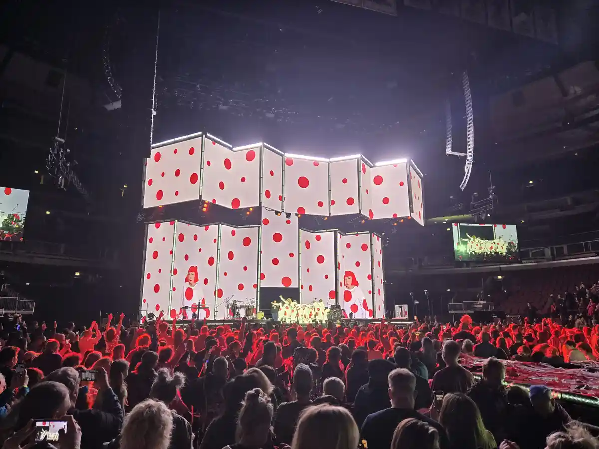 A wide shot of a concert venue filled with an enthusiastic crowd, looking up at a massive, multi-tiered stage structure…