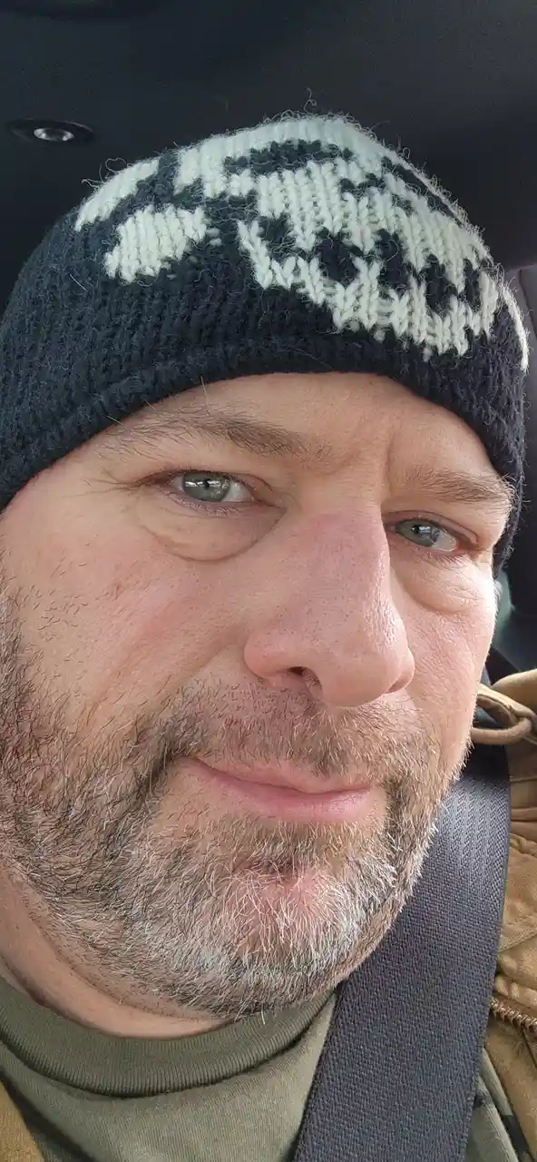 A close-up selfie of a middle-aged man with a graying beard and green eyes, wearing a dark gray knit beanie with a white…