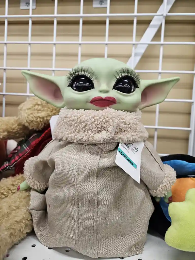 A green, baby Yoda plush toy is displayed on a store shelf, wearing a beige jacket with a fluffy collar and sporting…