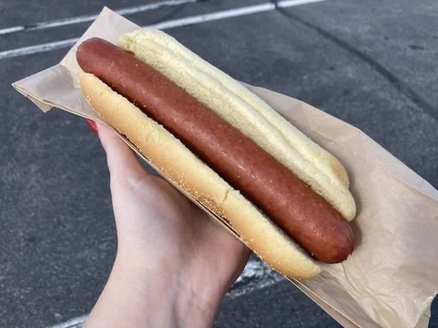 A hand holds a plain hot dog in a bun wrapped in brown paper, with a second, identical hot dog placed directly on top of it…