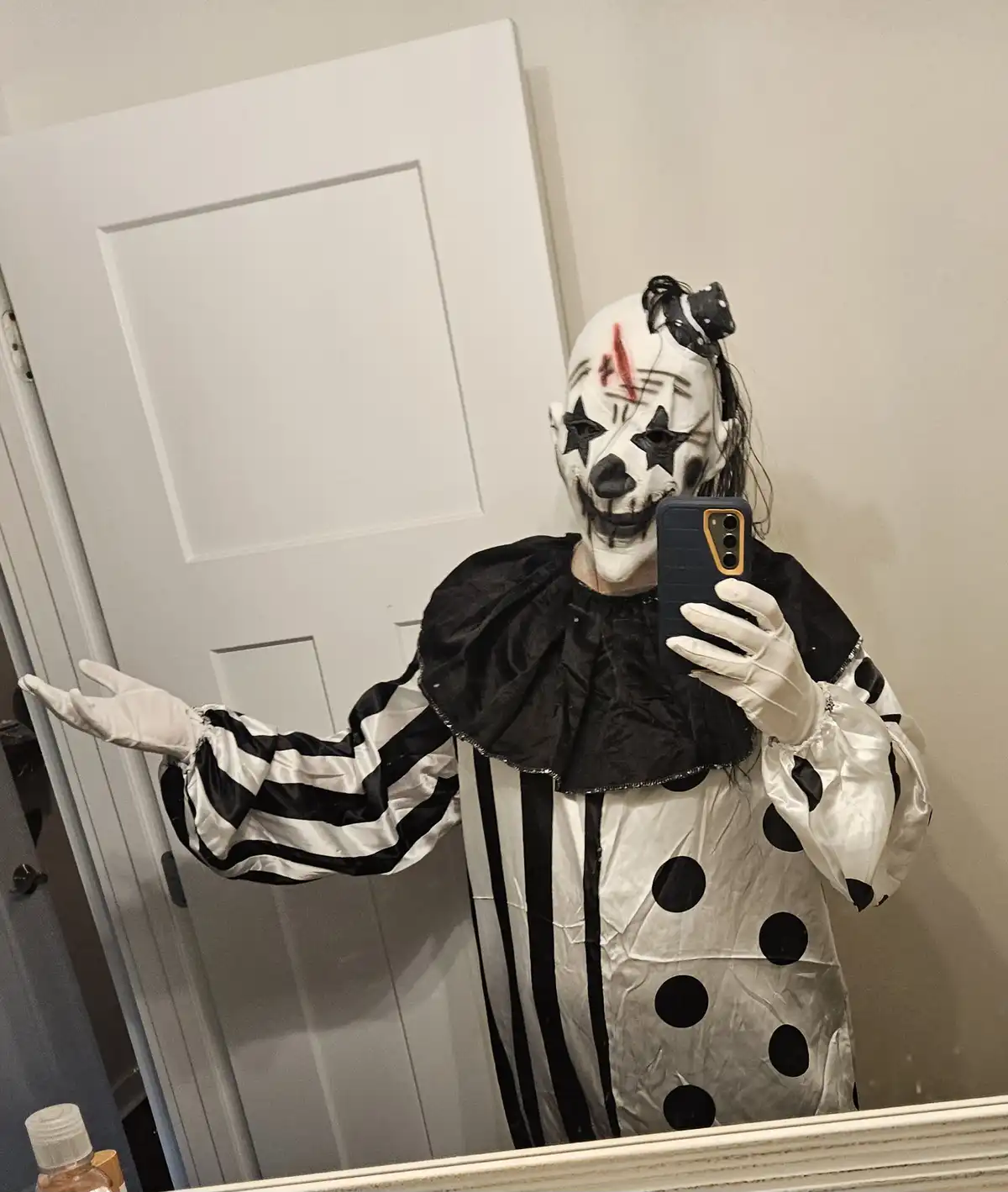 A person in a full clown costume with a white face painted with black stars, a red slash, and a black smile, wearing a…