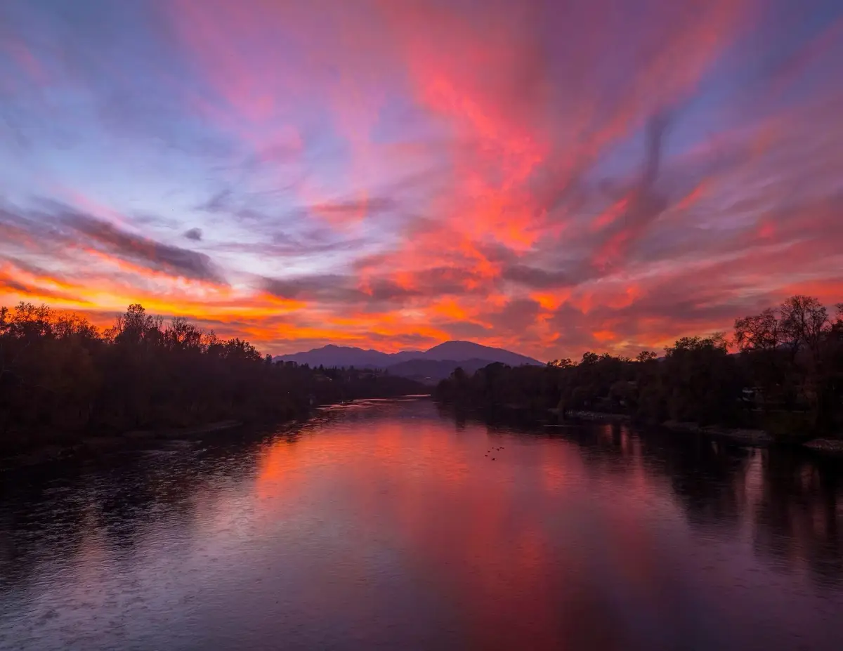 A serene landscape photograph shows a wide river reflecting a dramatic, colorful sky at sunset or sunrise, with vivid pink…