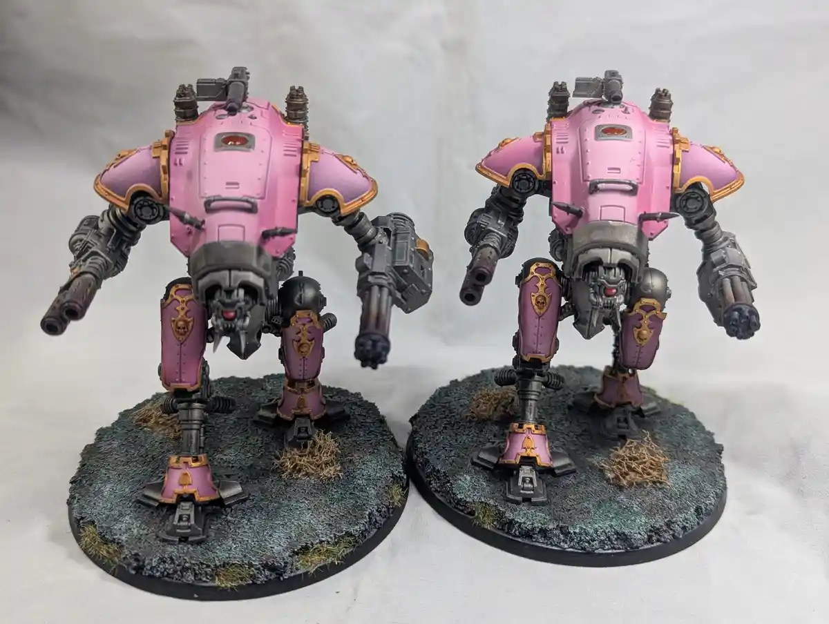 Two heavily armored, pink and gold-painted war machines stand side-by-side on textured bases against a white backdrop. The…