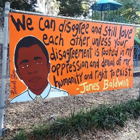 An orange painted sign on a chain-link fence features a cartoon portrait of Barack Obama and a quote attributed to James…