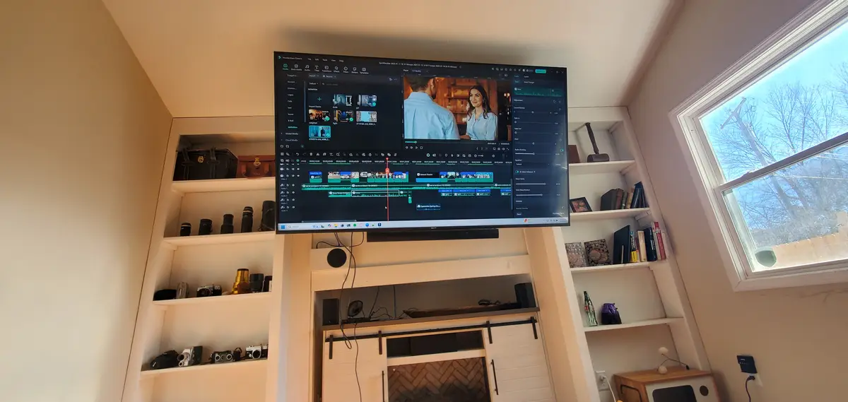 A large TV screen mounted above a fireplace displays a video editing software interface with clips and audio tracks, while…