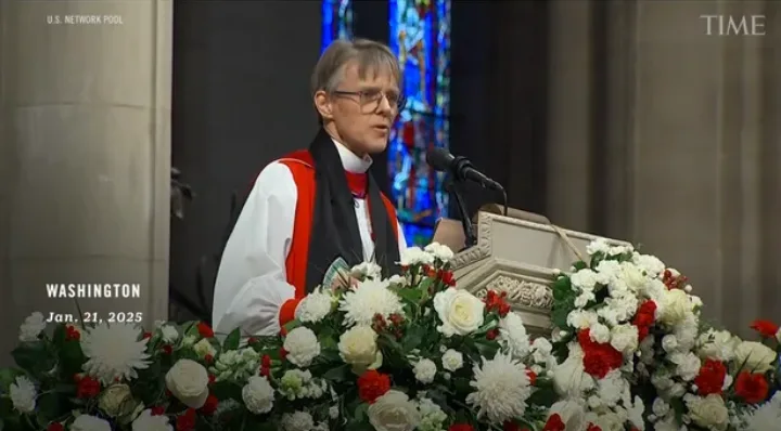 A woman in clerical attire, including a white shirt, black scarf, and red stole, stands at a floral-adorned podium in what…