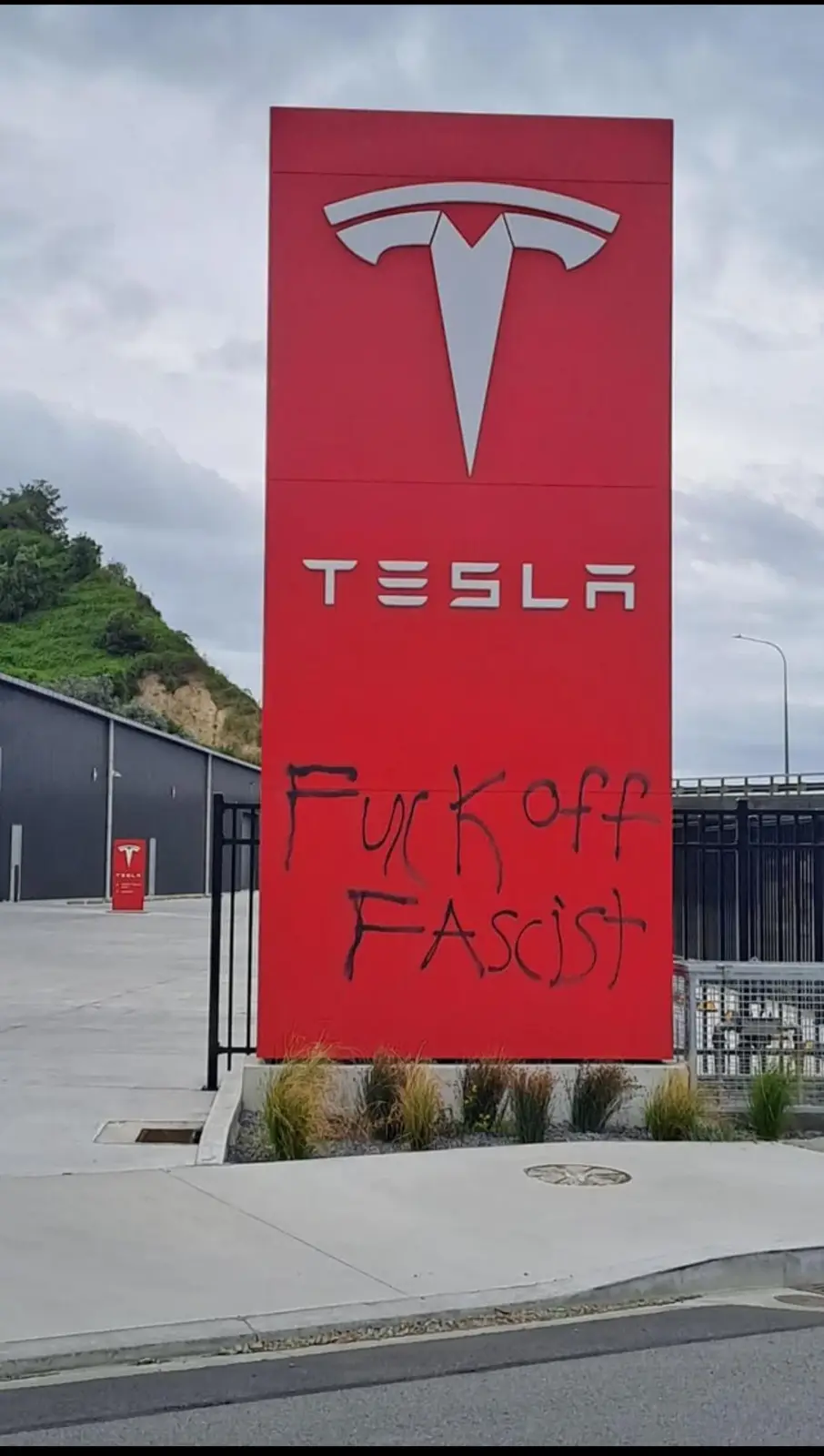 A large red Tesla sign with the company logo and name is visibly defaced with black spray-painted graffiti reading 'Fuck off…