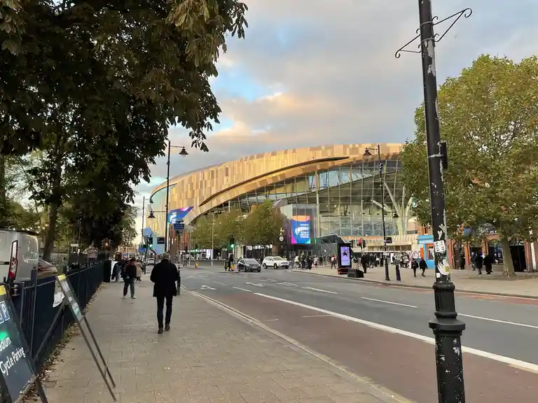 A street view of the exterior of Tottenham Hotspur Stadium, with its distinctive curved golden facade and glass structure…