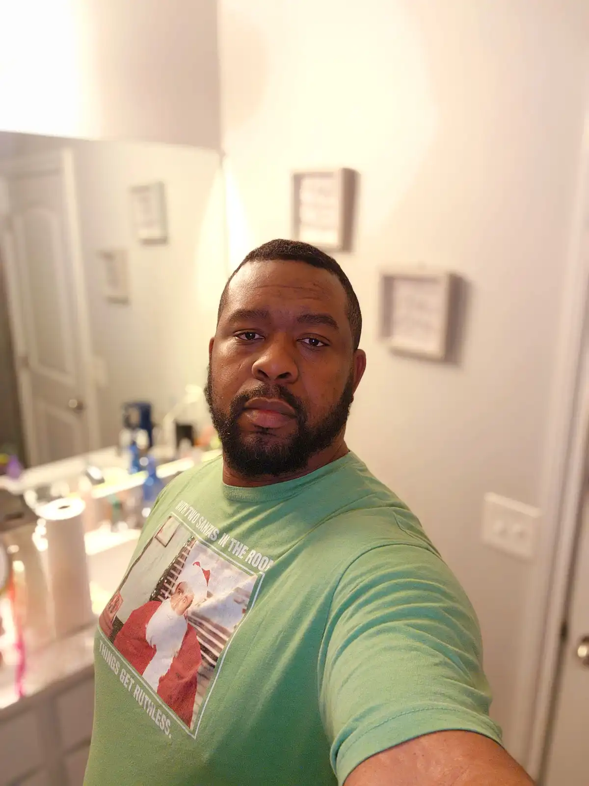 A man with a beard takes a selfie in a bathroom wearing a green t-shirt that features a graphic of a man in a Santa suit and…