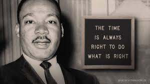 A black and white photo of Martin Luther King Jr. looking forward, with a framed sign beside him displaying the text 'THE…