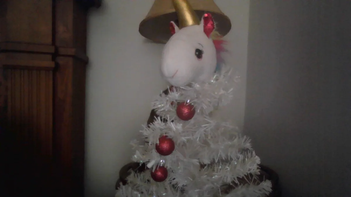 A plush white unicorn with a gold horn and red ears is dressed as a Christmas tree, wearing a gold lampshade hat and…