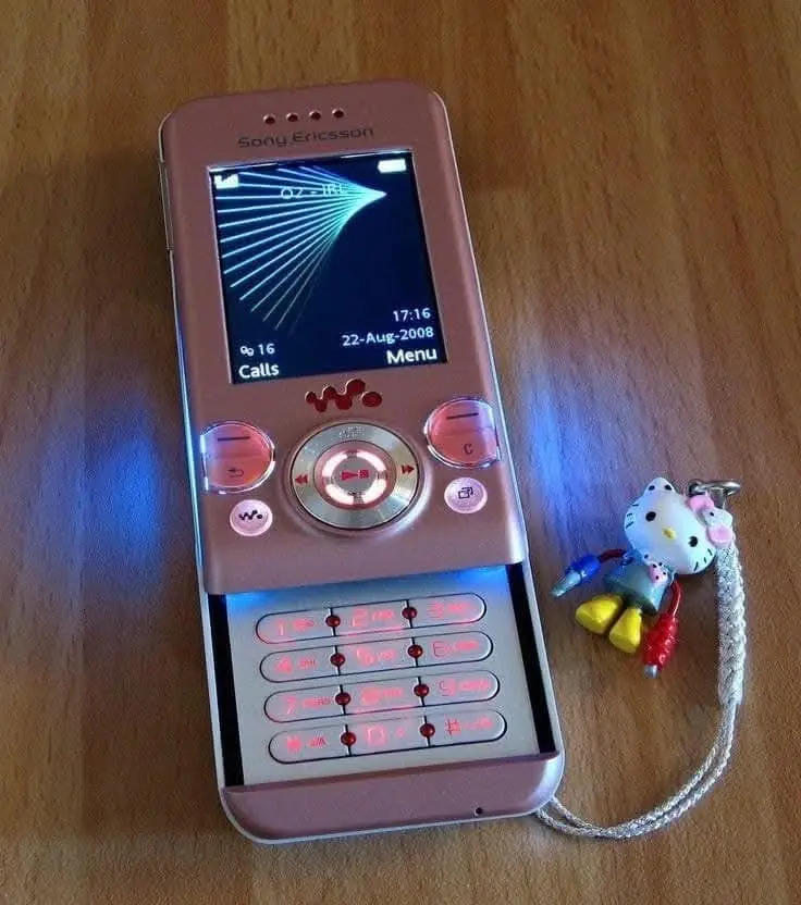 A pink Sony Ericsson W810i phone from 2008 with its screen on, displaying the time and date, sits on a wooden surface next…
