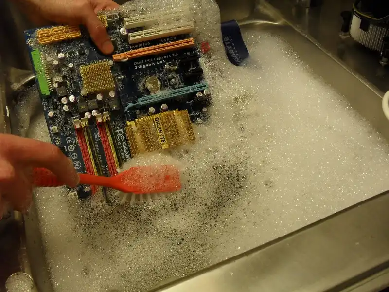 A person is scrubbing a computer motherboard with an orange toothbrush in a sink full of soapy water, with visible foam and…