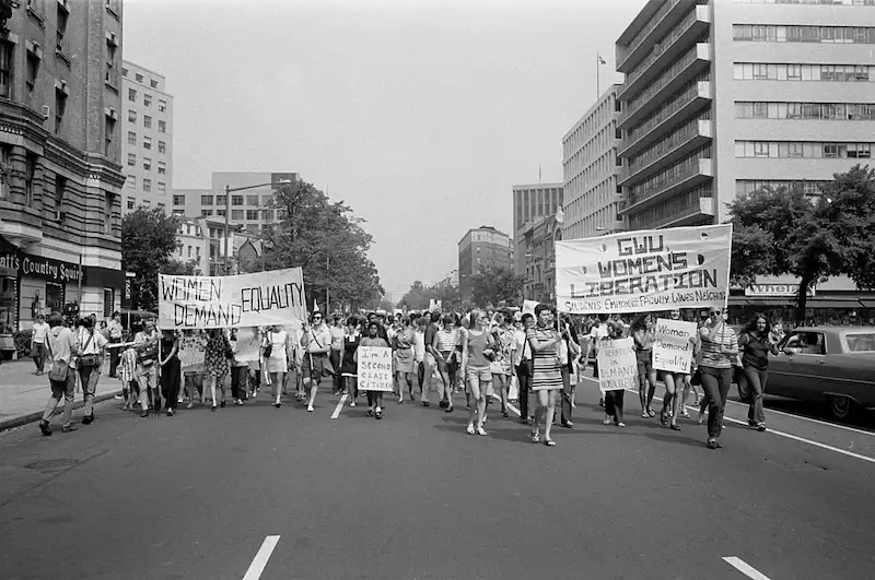 A black-and-white photograph of a 1970s women's liberation march in an urban street, with participants holding signs…