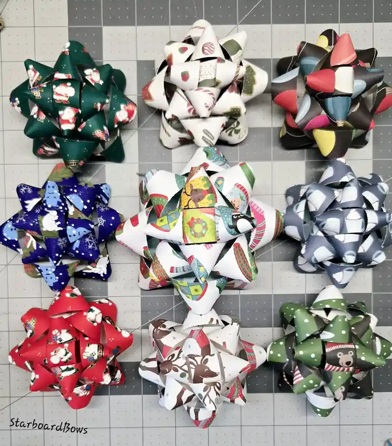 An overhead shot displays nine intricately folded paper bows arranged in a 3x3 grid on a gray grid-patterned cutting mat…