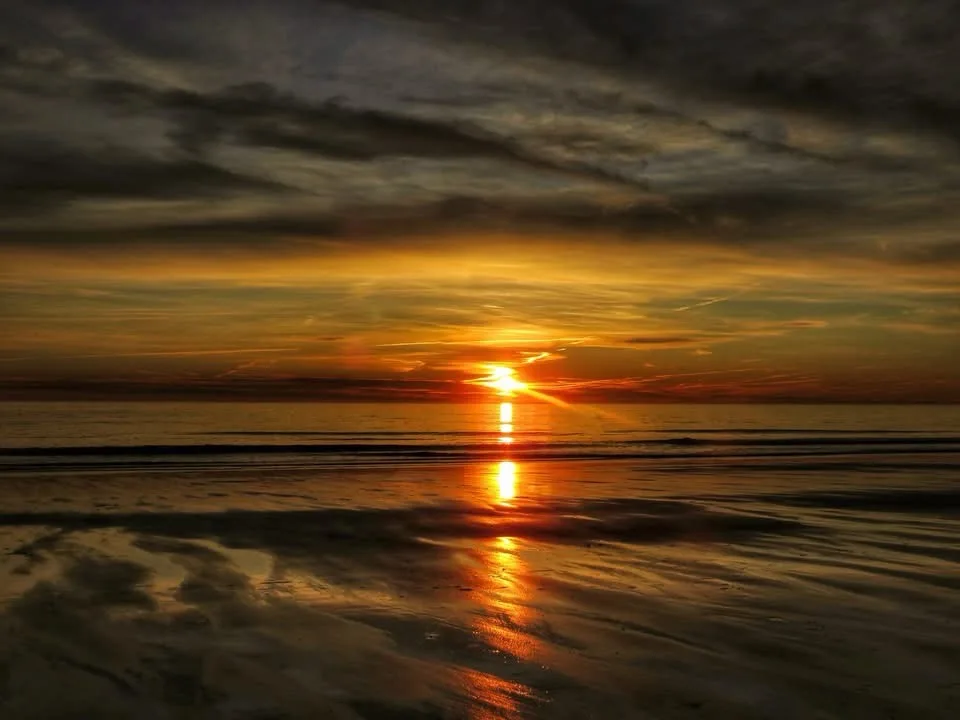 A serene sunset over a beach with the sun low on the horizon, casting golden light across the water and wet sand. Dark…