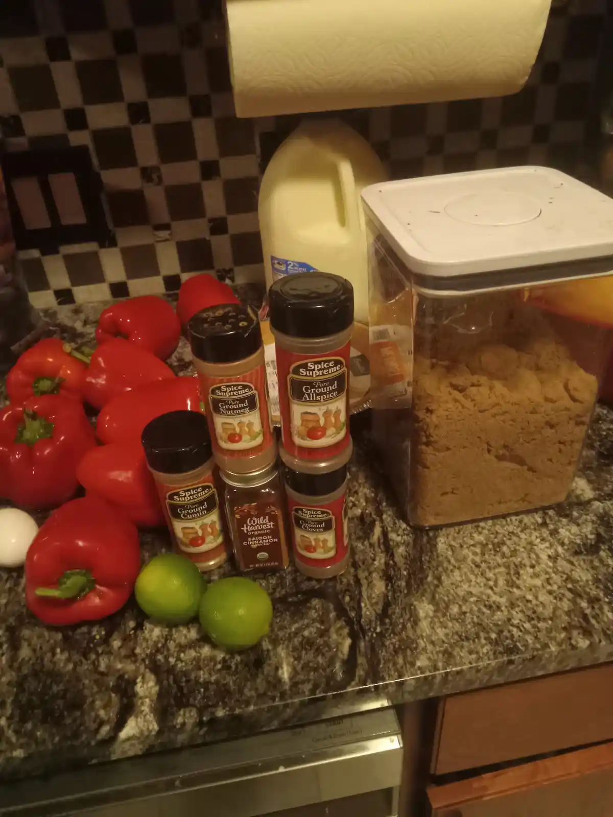 A kitchen counter displays a large pile of red bell peppers, two limes, an egg, and several spice containers including…