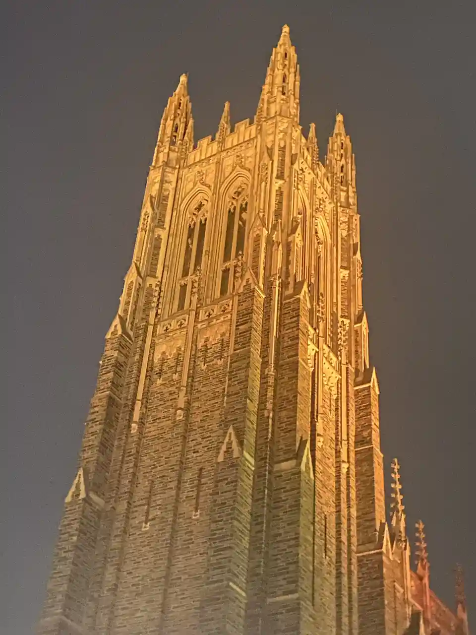 A low-angle nighttime photo shows a tall, ornate Gothic-style stone tower illuminated by warm yellow lights against a dark…