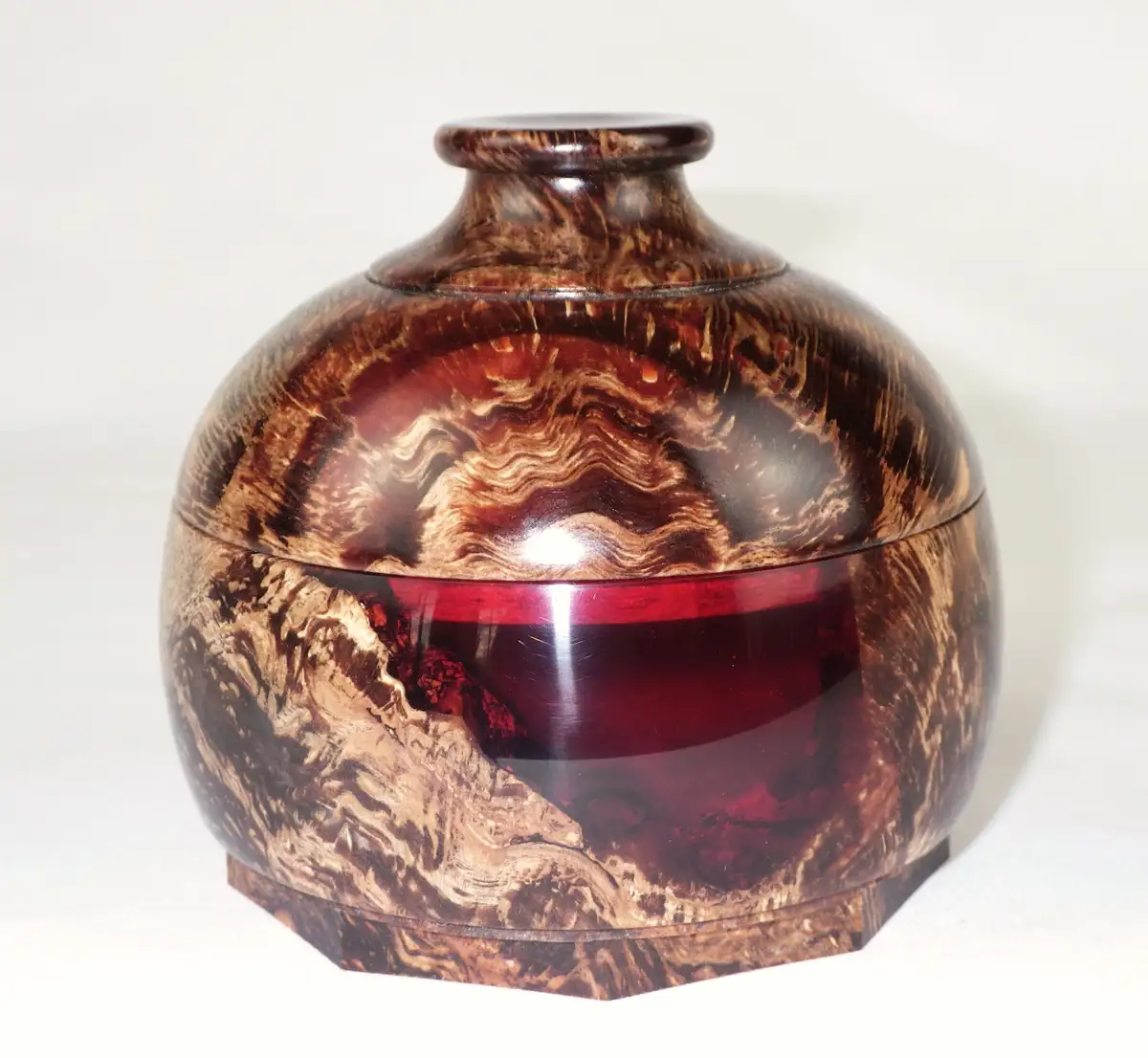 a polished wooden box with swirling brown and amber grain patterns, featuring a glossy red glass band around its middle, set…