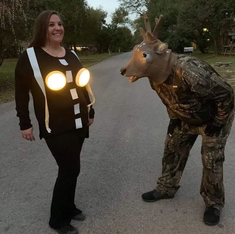 A woman in a black costume with glowing circular lights and white reflective stripes stands on a road, smiling. Beside her…