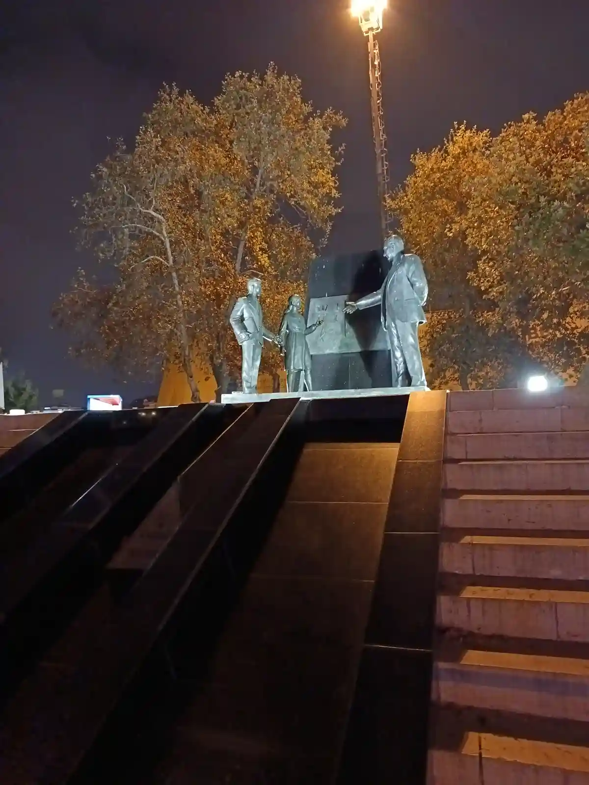 A nighttime photo of a bronze statue depicting three figures: a man on the right gesturing toward a plaque, a woman in the…