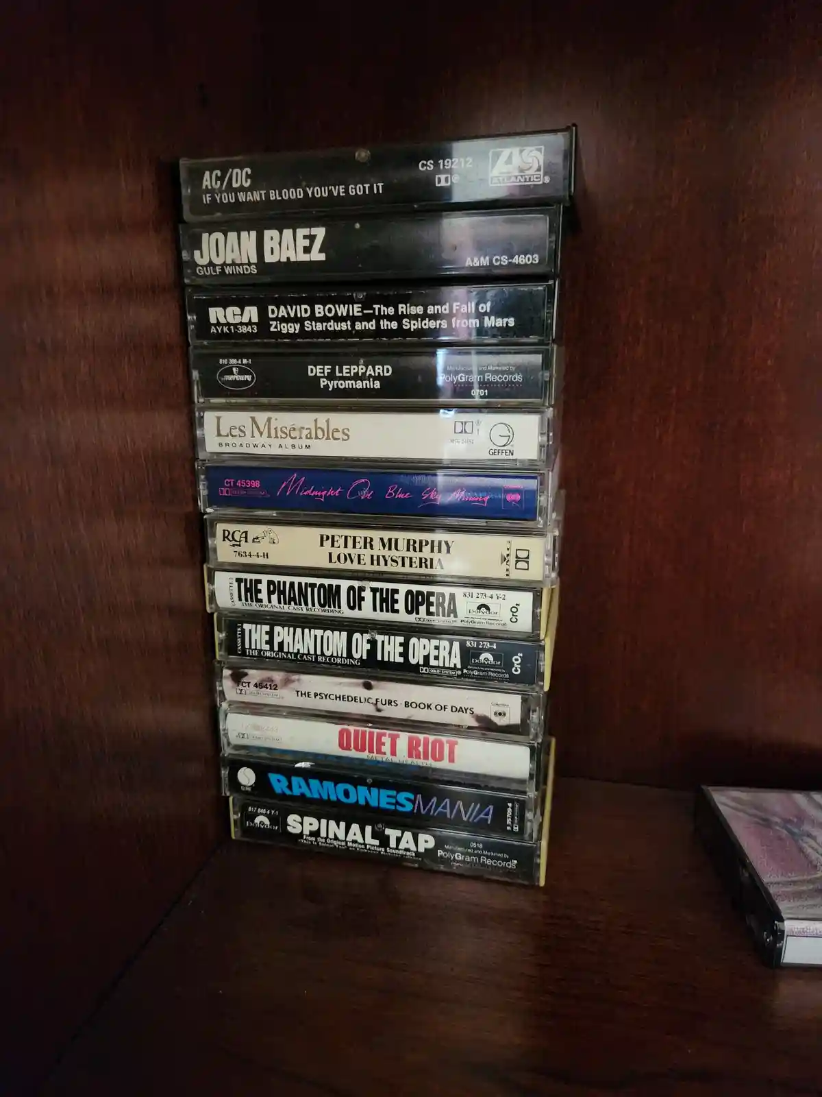 A vertical stack of cassette tapes sits on a wooden shelf, featuring artists like AC/DC, Joan Baez, David Bowie, Def…