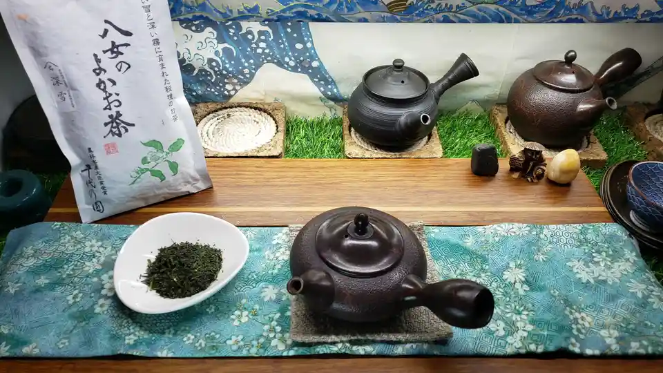 A still life of a traditional Japanese tea setup featuring three dark ceramic teapots on wooden trays, a white bowl of loose…