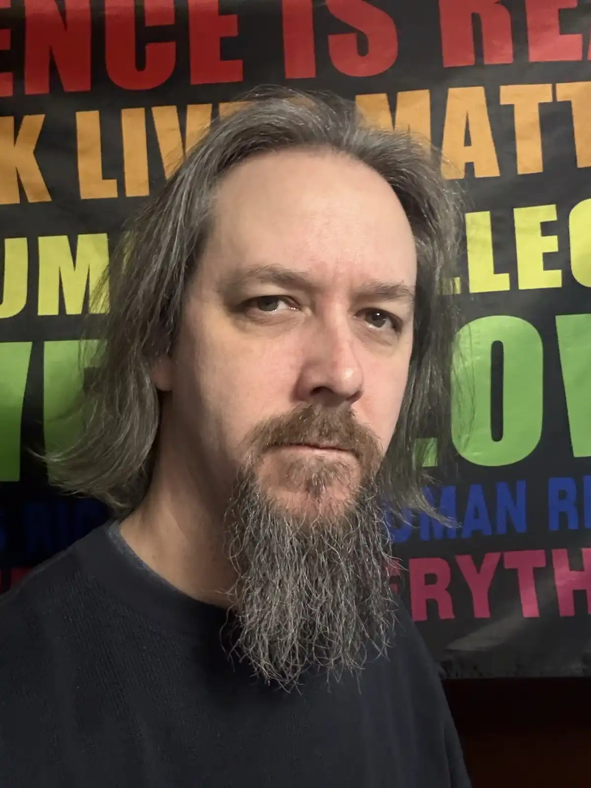 A man with long gray hair and a beard stands in front of a colorful protest banner with phrases like 'BLACK LIVES MATTER'…