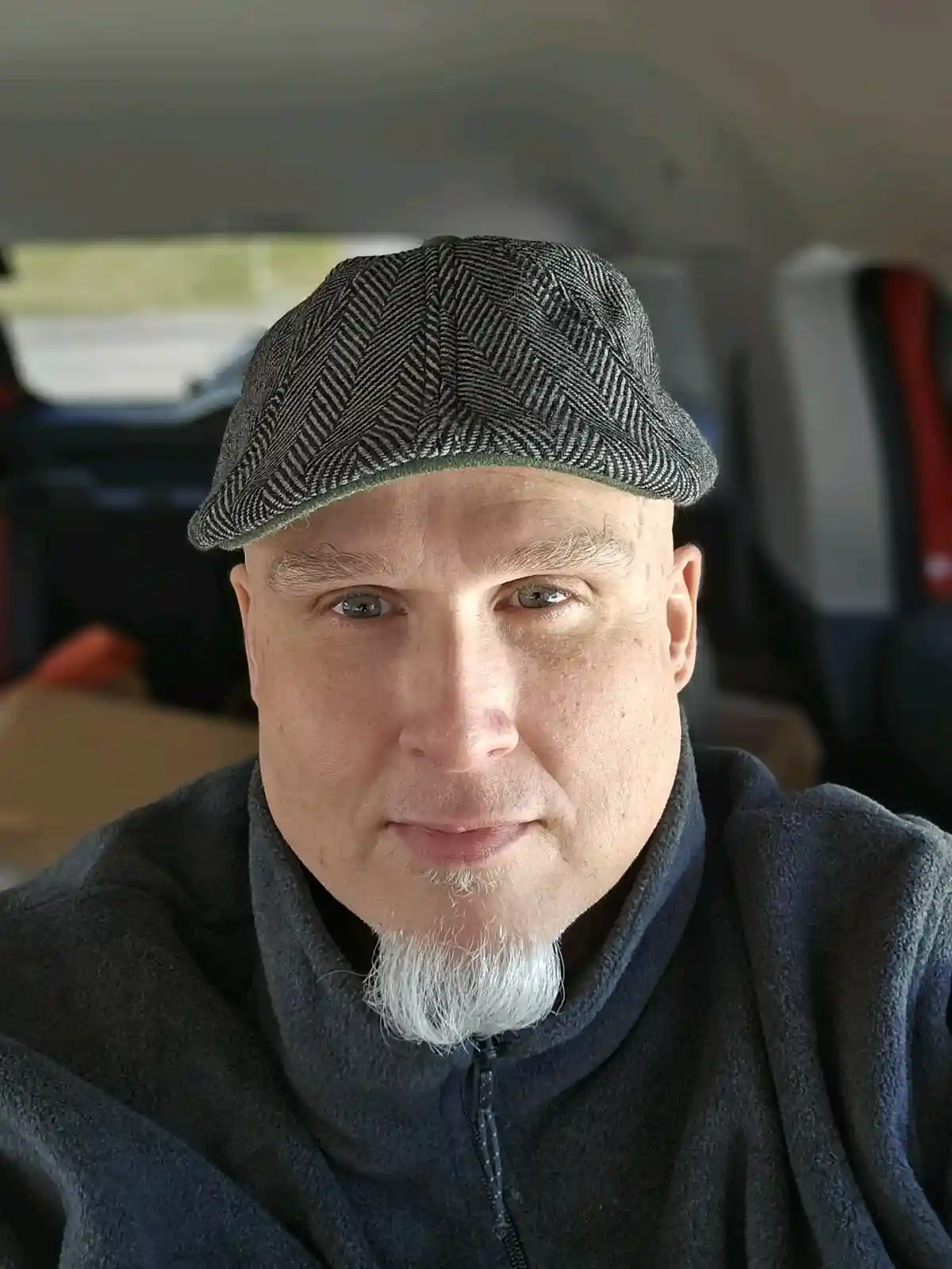 A bald man with a white goatee and a herringbone-patterned flat cap sits inside a car, looking directly at the camera with a…