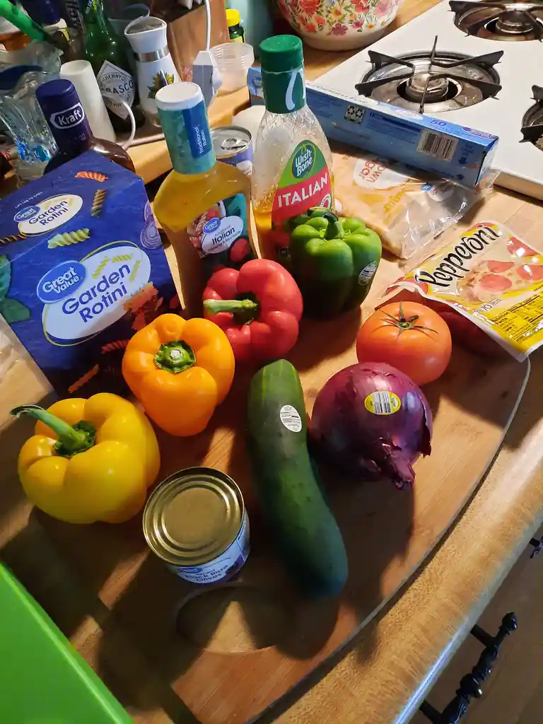 A cluttered kitchen counter displays a large assortment of ingredients including colorful bell peppers, a cucumber, a red…