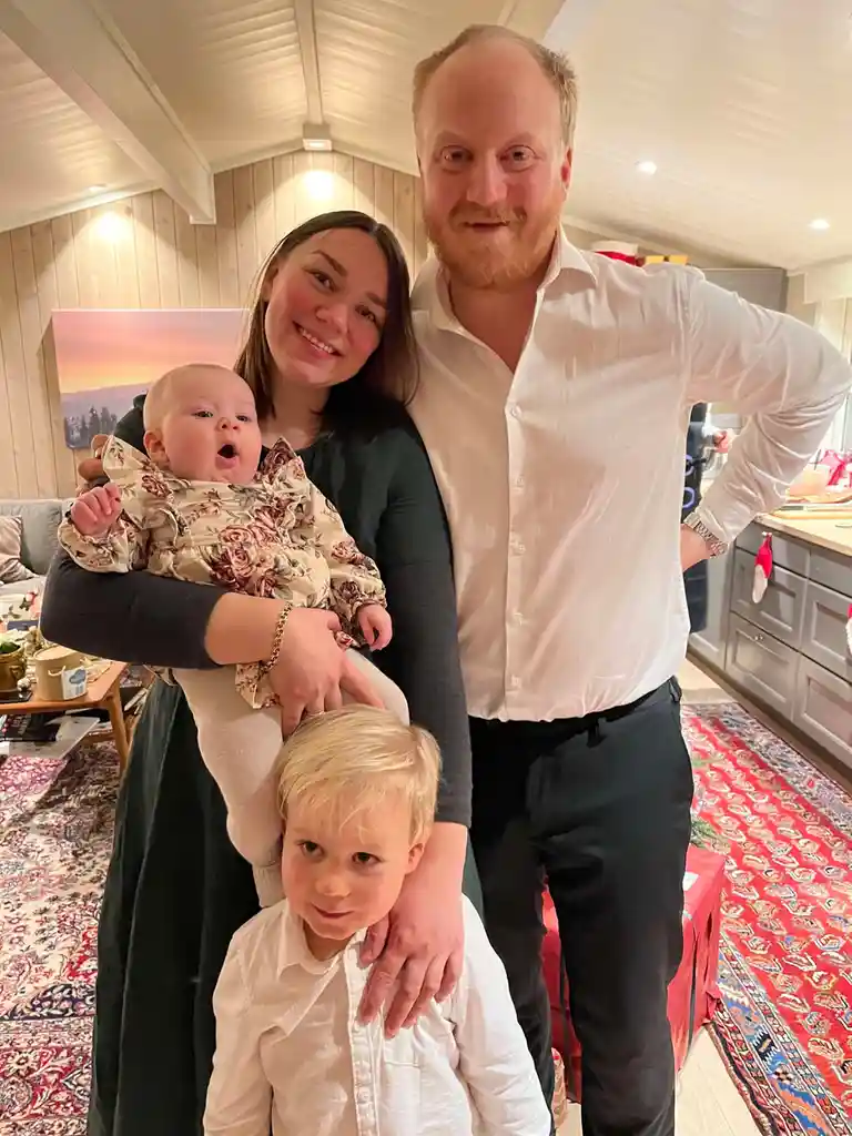A family photo shows a man, woman, and two children posing indoors. The woman holds a baby in a floral outfit who has its…
