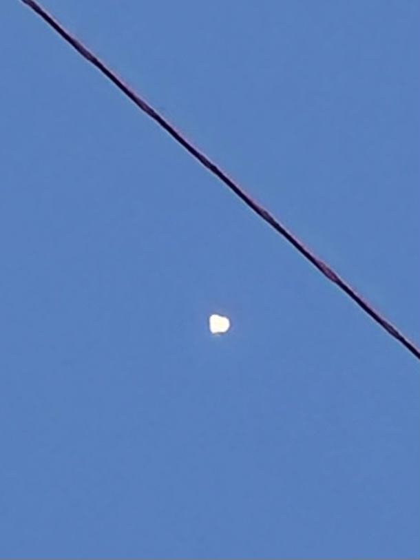 A clear blue sky with a diagonal power line stretching across the frame. Below the line, the moon appears as a bright…