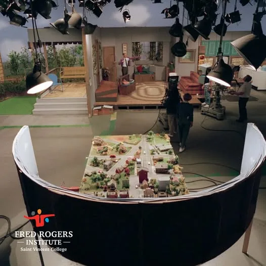 An overhead view of a television studio set, showing crew members and cameras filming a staged living room scene. In the…