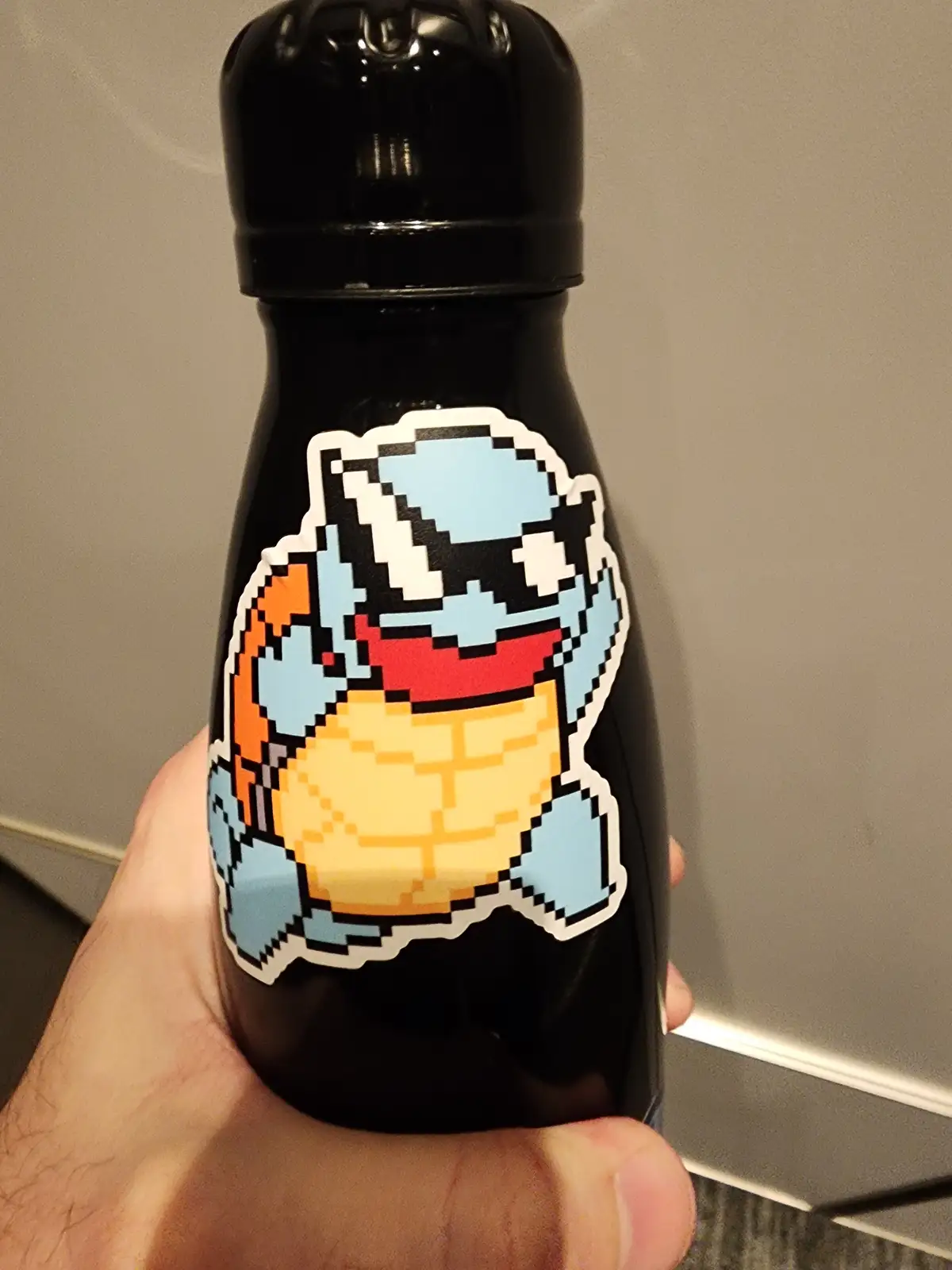 A hand holds a black water bottle with a pixelated sticker of the Pokémon Squirtle, which is depicted with a wide, toothy…