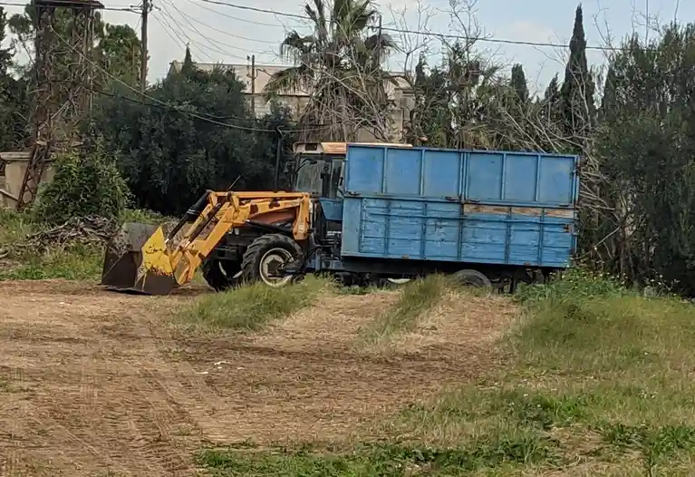 A yellow front loader tractor with a blue trailer is parked on a dirt path surrounded by grass and trees. The overlaid text…