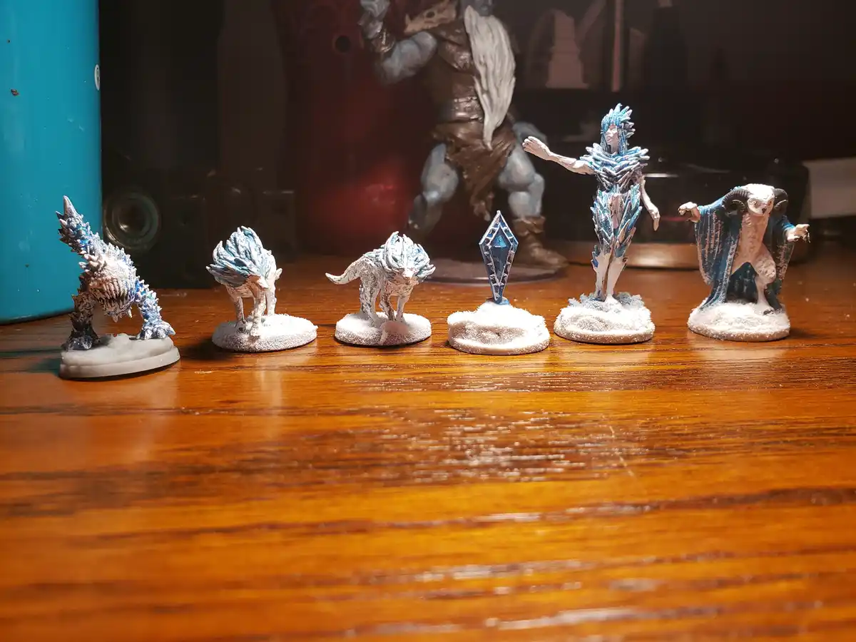 Six painted miniature figurines, mostly in blue and white to resemble ice or snow, are arranged in a row on a wooden…