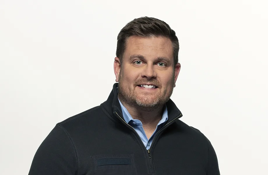 A man with short brown hair and a beard, wearing a dark zip-up sweater over a light blue collared shirt, smiles slightly…