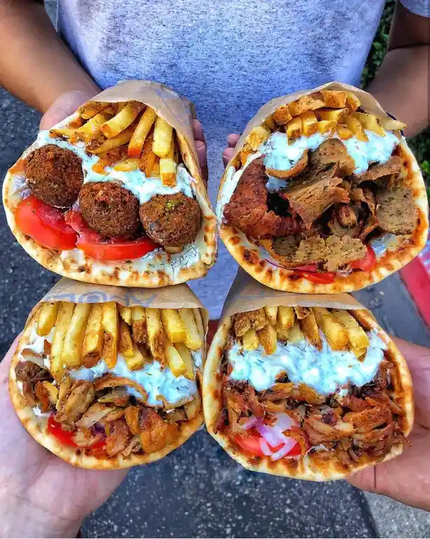 Four hands hold up four large, open-faced gyros wrapped in paper, each filled with different meats and toppings including…