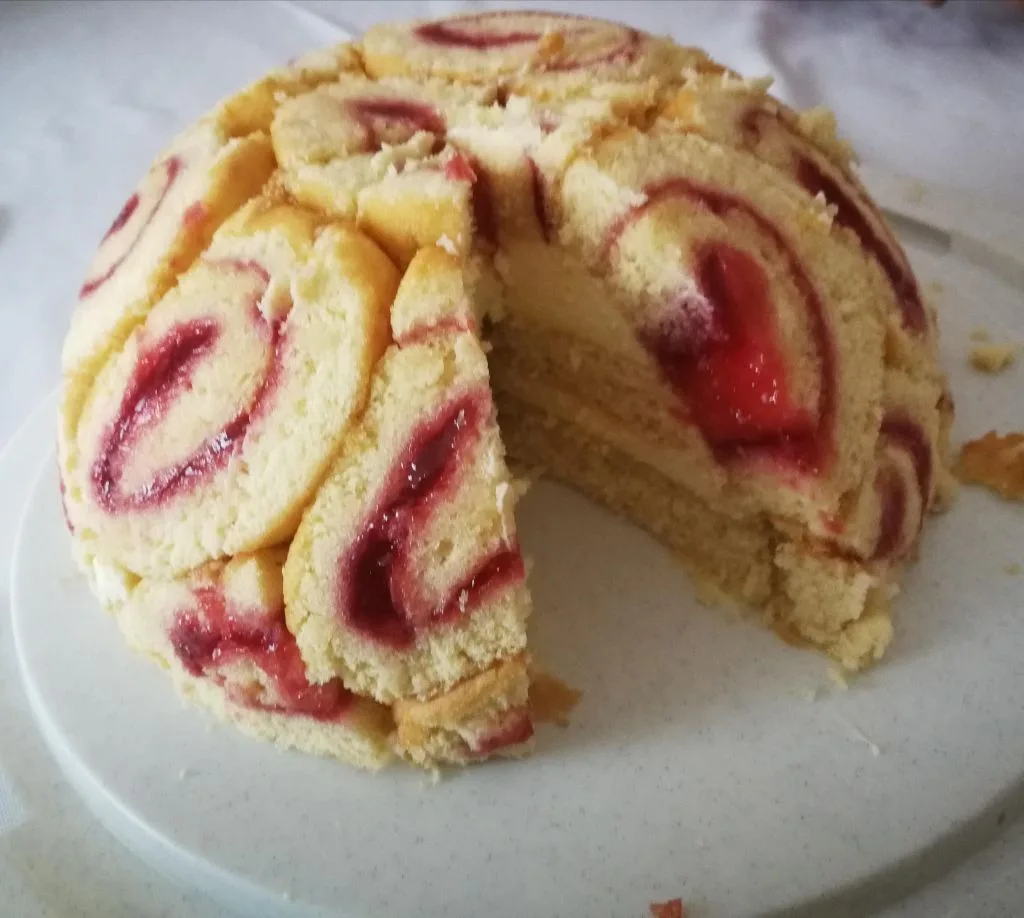 A close-up photo of a round, sliced cake with a pale yellow sponge and red swirls of jam or jelly inside, sitting on a white…