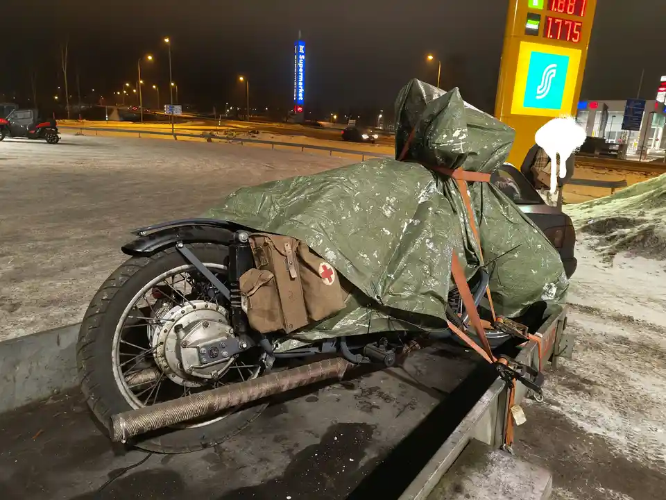 A motorcycle is heavily covered with a green tarp and strapped down on a trailer at a snowy gas station at night. A brown…