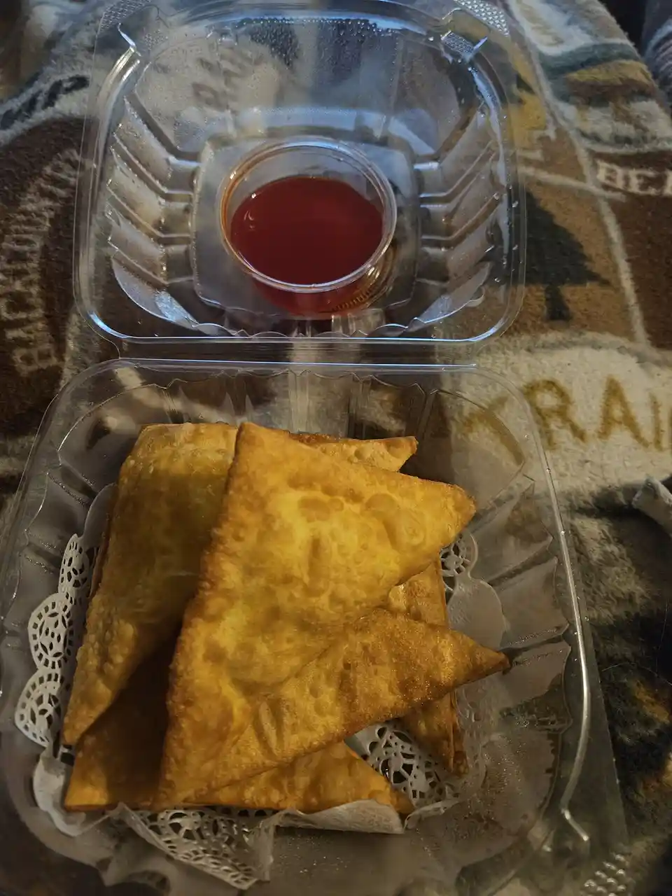 A clear plastic takeout container holds several golden-brown, triangular samosas resting on a white paper doily, with a…