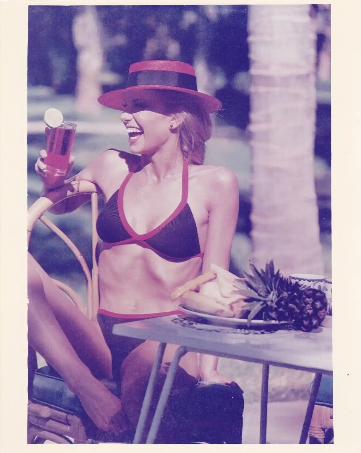 A woman in a black and red bikini and matching hat laughs while holding a drink with a lime wedge, seated at a table with a…
