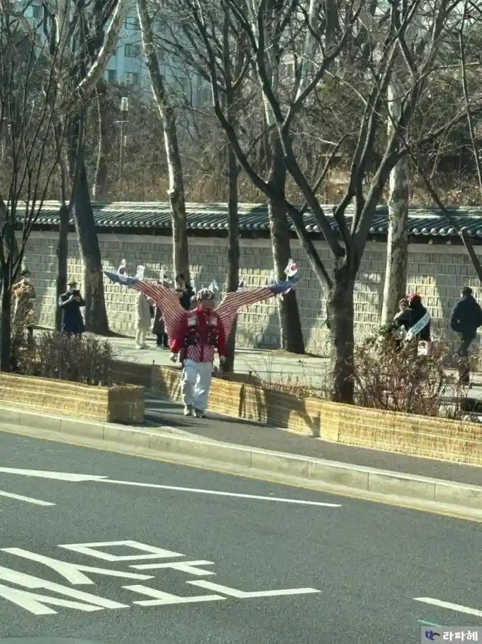 사람이 미국 국기 패턴의 날개를 뒤로 펼쳐서 걷고 있으며, 그의 옷은 붉은색과 흰색 줄무늬가 있고, 흰색 바지와 모자를 쓰고 있다. 배경에는 나무와 벽돌 벽, 그리고 다른 사람들이 보이며, 도로에는 한국어로 된 도로 표시가…