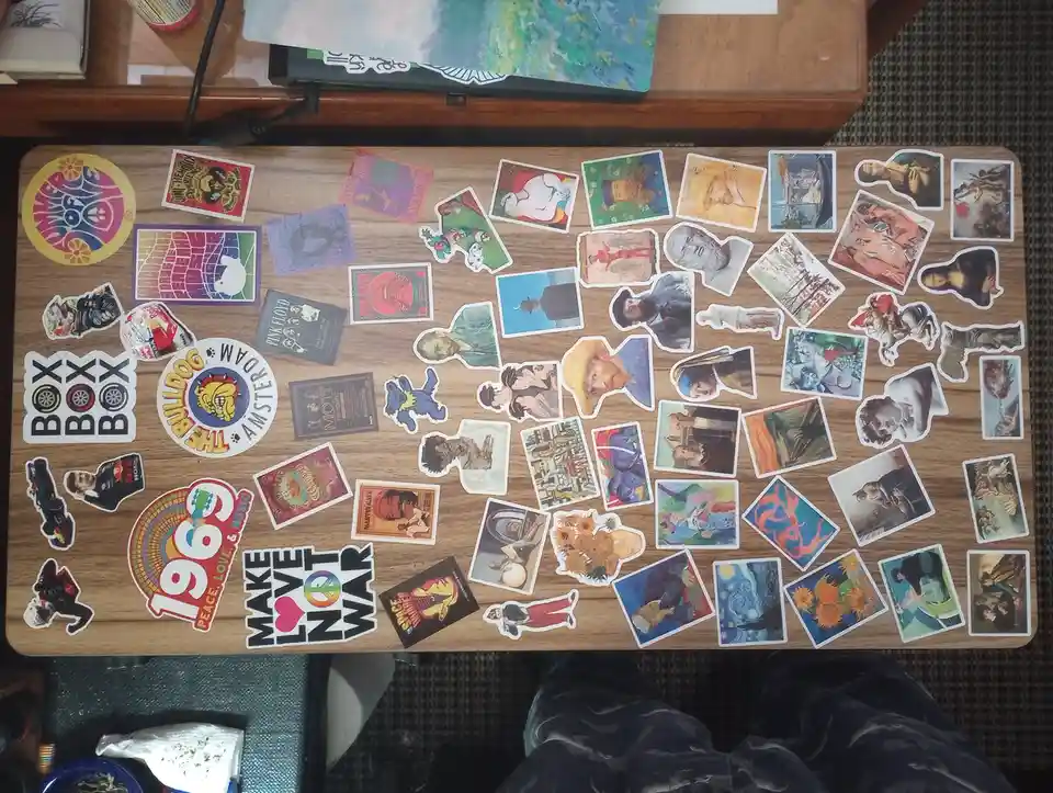 An overhead shot of a wooden table covered in dozens of stickers, including famous artworks like Van Gogh's sunflowers and…