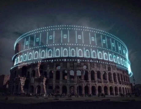 The ancient Colosseum in Rome is illuminated at night with bright blue lights arranged to form the word 'colosseum' in large…