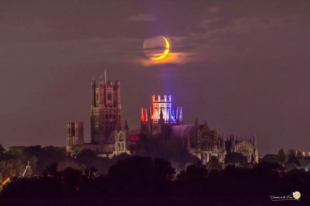 A night photograph shows Ely Cathedral in England, illuminated with red and blue lights, under a sky where a bright crescent…