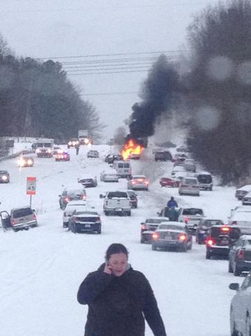 A woman in a dark jacket walks through a snowy, congested road with cars stopped in traffic. In the distance, a vehicle is…