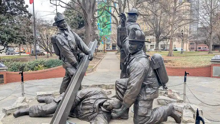A bronze statue depicts three firefighters in full gear; one kneels beside a fallen comrade, another stands holding a steel…