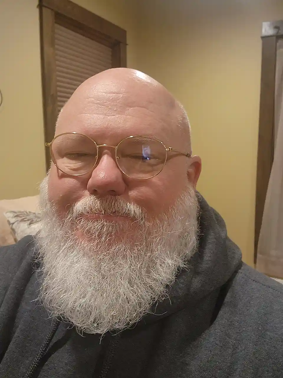 A close-up selfie of a bald man with a full white beard and gold-rimmed glasses, smiling slightly while wearing a dark grey…